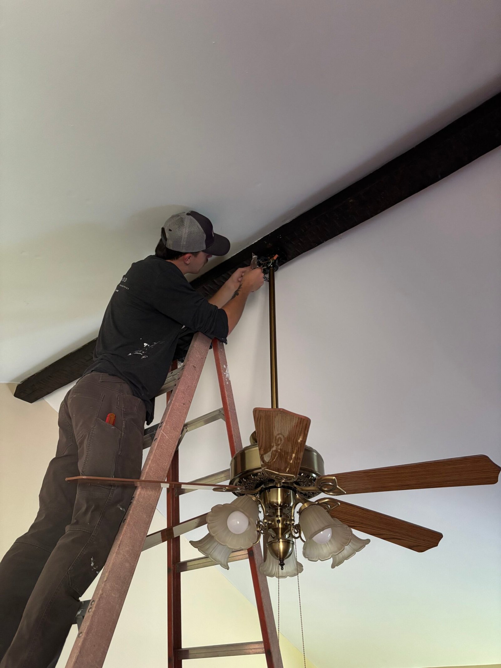Electrician on a ladder installing a ceiling fan and light fixture in a home with exposed beams